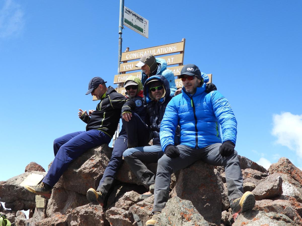 Trekkers celebrating at Point Lenana summit sign on Mount Kenya wearing full layered gear — guided by Ahambi Tours